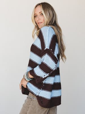 An oversized striped knit sweater in sky blue and deep mocha worn with relaxed jeans — the kind of cozy-cool piece that looks equally at home on a coffee run or curled up somewhere with good light.