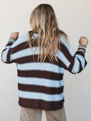 Shot from behind, a model in a chunky striped knit sweater in mocha and sky blue layers on stacked beaded bracelets — the kind of effortless, collected look that feels like it belongs on a weekend farmer's market run.