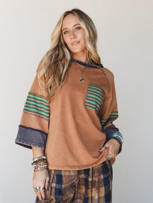 A relaxed, lived-in camel sweatshirt top with mixed pattern sleeves and a chest pocket — layered with stacked bracelets and a gold pendant necklace, it's the kind of outfit that looks effortless but very much on purpose.