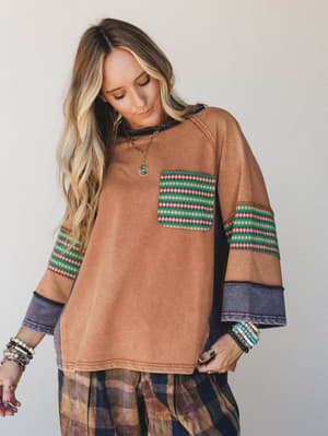 A relaxed, color-blocked pullover top in washed camel with contrasting navy panels and geometric diamond-print trim on the chest pocket and sleeves, styled with a plaid bottom and layered boho jewelry for a lived-in, expressive look.