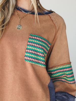 A close-up detail shot of a camel-toned sweatshirt with a diamond-print chest pocket and matching sleeve band, layered over a navy underlayer — the kind of top that looks like you've had it forever in the best way.