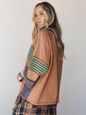 A relaxed oversized top in washed camel with contrasting zigzag-stripe trim at the sleeves and a denim-toned pocket, styled over a plaid midi skirt — the kind of piece that looks like it's been loved for years and fits like it knows you.