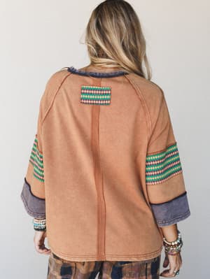 A relaxed oversized sweatshirt-style top shot from the back, done in washed camel with contrast diamond-print woven trim on the sleeves and a matching back patch — stacked bracelets on both wrists add that lived-in boho personality.