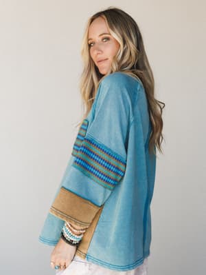 A relaxed oversized top in faded teal with a tribal-stripe sleeve panel and contrast camel cuff — the kind of piece that looks intentional with stacked bracelets and zero effort.
