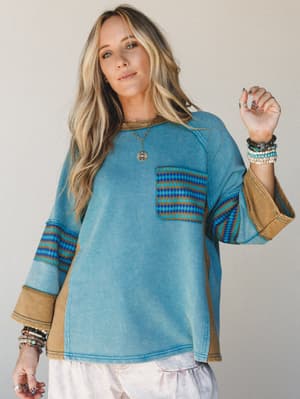 A faded teal oversized sweatshirt top with warm camel contrast cuffs and a geometric Aztec-print chest pocket, styled with stacked bracelets and a pendant necklace — casual boho with just enough personality to make it interesting.