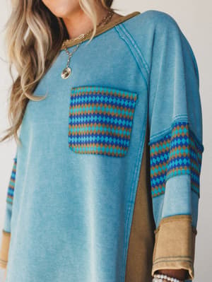 A close-up of a faded teal sweatshirt-style top with warm olive contrast paneling and a bold diamond-print chest pocket — the kind of top that looks like it's been loved for years and somehow only gets better.
