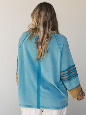 A relaxed, oversized top photographed from the back, showing off its faded teal washed fabric, contrast tribal-stripe sleeve panels in teal, gold, and navy, and a center back seam detail — the kind of piece that looks like it has a good story.