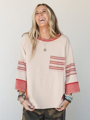 A laughing model in a relaxed, oversized latte-toned sweatshirt top with contrast red ringer trim and diamond-print sleeve and pocket detail — styled with camo pants and stacked boho bracelets for a lived-in, effortlessly cool look.