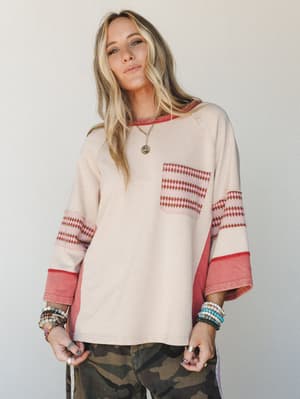 An oversized cream sweatshirt top with diamond-print contrast panels and rust-red cuffs, styled with camo pants and layered boho jewelry — the kind of top that looks like you tried just enough.