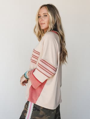 A side-profile studio shot of a relaxed oversized top in warm cream with dusty rose colorblock panels and geometric stripe trim at the sleeves and chest pocket, styled with camo pants and layered boho bracelets for an effortlessly eclectic everyday l
