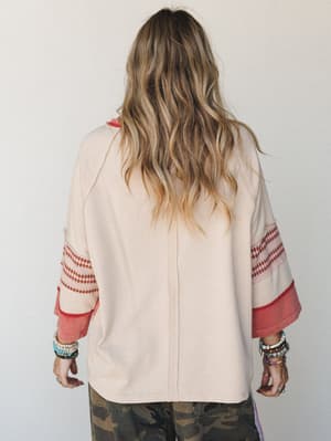 Shot from behind, this oversized cream top has serious personality — wide bell sleeves with a diamond-patterned fair isle band and a layered coral cuff give it that effortless boho-vintage feel, styled with stacked bracelets and camo pants for a look