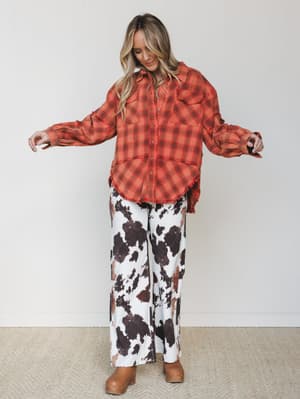 A model shows off a bold mix-and-match moment — an oversized rust plaid flannel with frayed hem edges paired with wide-leg cow print pants and tan platform clogs, arms outstretched like she's about to take the whole look for a spin.