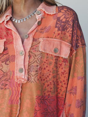 A close-up of a patchwork floral button-down blouse in warm tangerine and plum tones, styled with a delicate silver charm necklace — the frayed raw edges and mismatched prints give it that perfectly worn-in, found-in-a-vintage-market energy.
