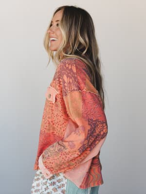 A side profile of a smiling model in a relaxed patchwork blouse with mixed floral and botanical prints in warm tangerine, coral, and plum tones — layered over a floral skirt, shot against a soft studio backdrop with an effortlessly boho feel.