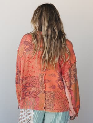 A back-facing model shows off a relaxed long-sleeve top in a warm tangerine patchwork print, with visible raw-edge seam detailing and a soft, lived-in feel — paired with teal striped wide-leg pants for a colorful boho mix.