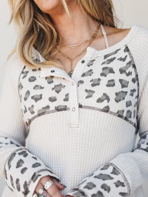 A cozy waffle-knit henley with leopard print color-blocking on the yoke and sleeves, styled with layered gold chain necklaces and stacked rings — the kind of top that looks intentional without trying.