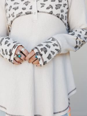 A close-up detail shot of a cream waffle-knit henley top with leopard print contrast sleeves and exposed stitch trim, styled with a stack of silver and turquoise rings that feel like they've been collected over years.