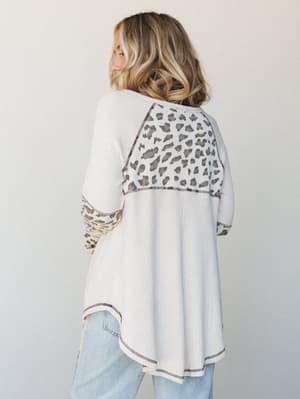 A back-view shot of a cream waffle-knit top with leopard print color-block panels at the yoke and sleeves, paired with light wash jeans — the kind of top that feels like staying in but looks like going out.