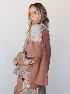 A model glances over her shoulder in a patchwork mixed-print top in warm mocha, layered over a flowing printed skirt — the kind of outfit that looks like it was put together effortlessly but definitely wasn't.