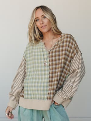 A mixed-pattern patchwork pullover in sage plaid and warm tan stripe gets styled with soft mint trousers and layered pendant jewelry — effortlessly relaxed with just enough personality to make it interesting.
