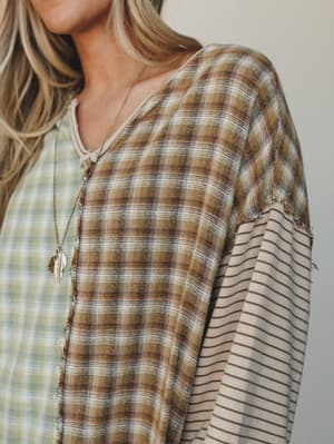 A close-up of a mixed-print long sleeve top in earthy sage and olive tones, featuring patchwork plaid and stripe panels with raw-edge seams — styled with a delicate feather pendant necklace for an effortlessly lived-in, boho feel.