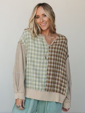 A mixed-pattern long sleeve top in sage plaid and earthy tan stripe steals the show — the kind of piece that looks like it wandered out of a vintage shop and into your weekend rotation, styled with soft mint pants and layered silver jewelry.