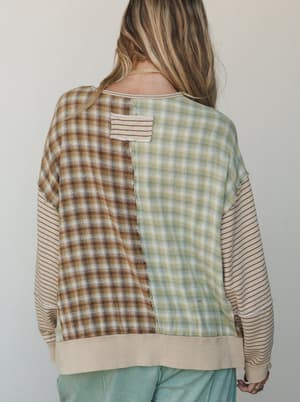 A back-view shot of a mixed-media pullover top with two contrasting plaid panels — one warm brown, one sage green — joined down the center back, with striped long sleeves and a ribbed hem, giving it that intentionally patchwork, lived-in feel.