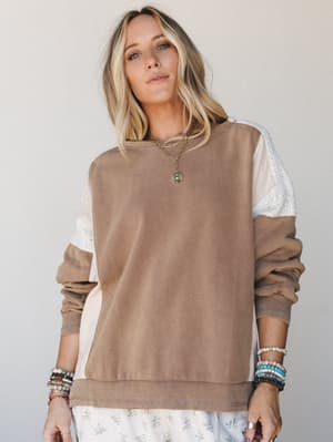 A relaxed color-block sweatshirt in mocha and cream with lace panel inserts worn over a floral skirt, layered with beaded bracelets and a gold pendant necklace — the kind of top that makes a Saturday feel intentional.