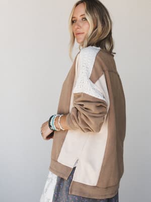 A relaxed oversized sweatshirt in mocha and cream colorblocking with lace panel detailing on the sleeve, styled over a floral skirt with a stack of beaded bracelets — the kind of top that looks like you tried just enough.