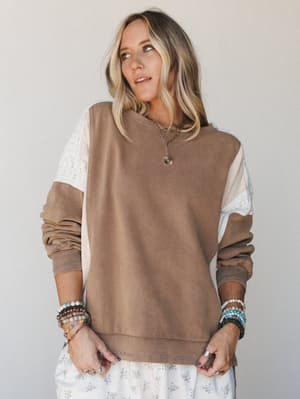 A relaxed colorblock sweatshirt in warm mocha with cream and lace panel sleeves, styled with a floral skirt and a stack of boho bracelets — the kind of top that makes a Tuesday feel intentional.