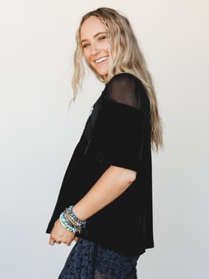 A smiling model shot in three-quarter profile wearing a relaxed black velvet top with sheer mesh shoulder panels, styled with stacked turquoise and beaded bracelets and a glimpse of dark printed bottoms — effortlessly boho with a warm, candid energy.
