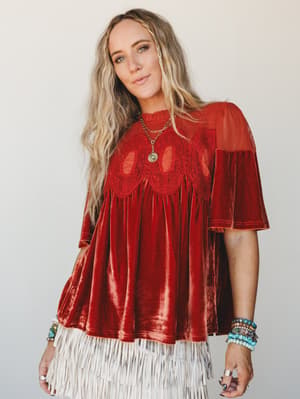 A rust velvet babydoll top with crochet chest detail and sheer mesh shoulders, styled with a cream fringe skirt and layered turquoise and mixed-stone bracelets — the kind of outfit that feels equal parts vintage and intentional.