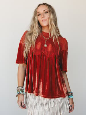 A velvet rust top with crochet lace chest detail and a flowy babydoll silhouette, styled with a cream fringe skirt and layers of beaded bracelets — the kind of outfit that looks like it was made for a bonfire and a good playlist.