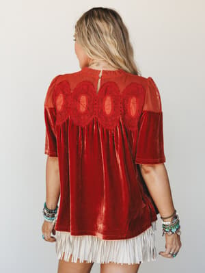 Shot from behind, a rust velvet babydoll top with a sheer mesh yoke and crochet appliqué detail takes center stage — the kind of piece that looks just as good walking away, styled with stacked boho bracelets and a white fringe skirt peeking out below