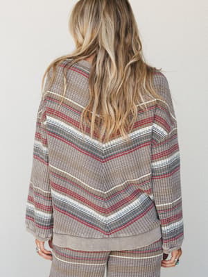 A back-view shot of a relaxed waffle-knit pullover in a warm ash base with multicolor horizontal stripes — the kind of top that looks equally at home on a slow Sunday morning or a casual coffee run.
