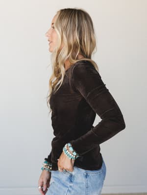 A side-profile shot of a model in a fitted dark brown velvet long-sleeve top paired with light wash jeans and a stack of turquoise and cream beaded bracelets — relaxed and effortlessly put-together.