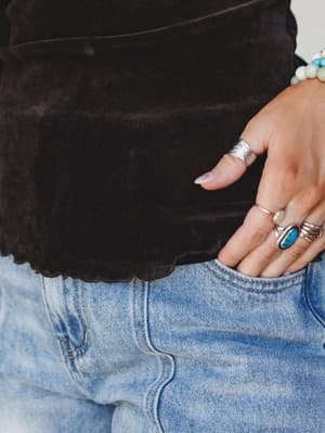 A close-up detail shot of a soft velvet top in deep charcoal tucked loosely into light-wash jeans, hand resting in pocket and stacked with silver rings and a turquoise stone — the kind of effortless layering that looks like it took zero effort and ex
