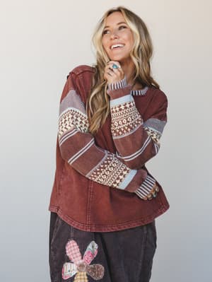 A laughing model in a washed mocha mixed-media top with Fair Isle knit sleeves and a curved hem, styled with dark corduroy pants featuring a patchwork floral appliqué — the whole look gives 'cozy Saturday with good light' energy.