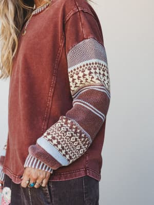 A close-up detail shot of a washed rust-red sweatshirt with mixed-media patchwork knit sleeves in Fair Isle and stripe patterns, styled with corduroy pants and stacked silver and turquoise rings — the kind of piece that looks like it's been loved for