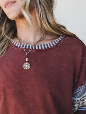 A close-up of a lived-in mineral-washed top with a striped contrast neckline and fair isle sleeve detail, styled with a layered gold chain and 'mama' coin pendant — the kind of outfit that feels personal without trying too hard.