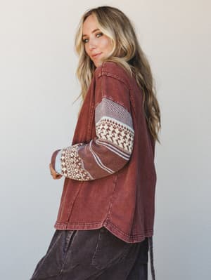 A blonde model glances back over her shoulder in a washed rust-mocha pullover top with contrast fair isle knit sleeves — the kind of relaxed, lived-in piece that feels like a weekend well spent.