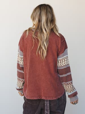 A back-view shot of a relaxed mixed-media top in washed brick red, with cozy fair isle knit sleeves and a soft corduroy bottom half — the kind of piece that makes a Saturday morning feel intentional.