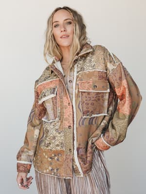 A relaxed, confident model wears an oversized patchwork floral shacket with sherpa trim — the kind of jacket that makes a whole outfit feel like a decision, styled over striped wide-leg pants in a clean studio setting.
