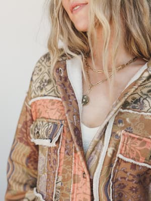 A close-up of a patchwork floral shacket with sherpa trim layered over a white tank, styled with gold layered necklaces — the kind of jacket that does all the talking.