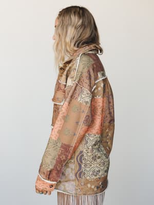 A side profile of a model wearing a patchwork floral shacket in warm earthy tones — tawny, blush, and dusty plum — with raw edge seam trim and fringe hem, shot clean against a soft studio wall.