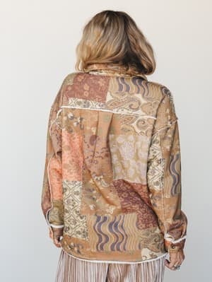 A back-view shot of a patchwork shacket in warm earthy tones — floral, paisley, and wave prints sewn together in a quilt-like layout with visible white fringe trim at the seams and hem, worn over a striped skirt in a clean studio setting.