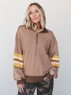 An oversized quarter-zip pullover in washed mocha brown gets a retro-sporty twist with mustard and white striped ribbed sleeve bands, styled casually with camo pants and silver stacked rings.