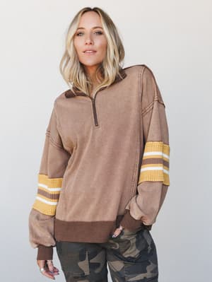 An oversized acid-wash quarter-zip pullover in mocha brown with retro varsity stripe cuffs, styled with camo pants for an effortlessly cool, lived-in look.