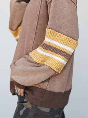 A close-up detail shot of a washed mocha sweatshirt with a chunky mustard yellow ribbed cuff trimmed in brown and white stripes — earthy, lived-in, and the kind of top that makes a camo jean feel intentional.