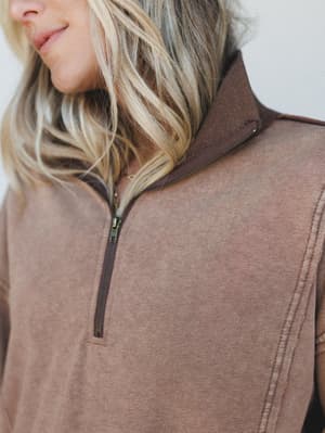 A close-up detail shot of a washed mocha quarter-zip pullover, highlighting the contrast zipper placket and tonal seam stitching — the kind of top that looks like you meant to look this good.